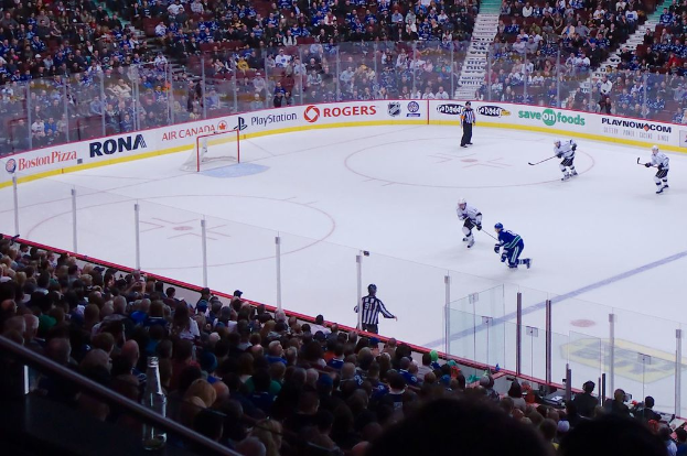 Vier MÃ¤nner beim Eishockey auf einem Eisstadion, mit zwei stehenden und zwei anderen in Bewegung, einem Tor und einem Zaun dahinter, Zuschauern auf StÃ¼hlen im Vorder- und Hintergrund, einem GelÃ¤nder und einer Treppe in der Ferne.