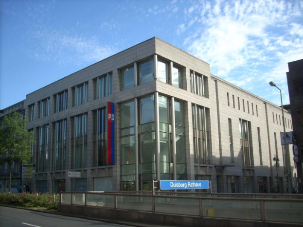 Ein großes Gebäude mit zahlreichen Fenstern, das als Hauptquartier der Deutschen Bank dient, umgeben von Straßenlaternen, Straßenschildern, Motorfahrzeugen, Büschen, Bäumen und einem bewölkten Himmel.