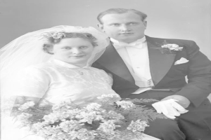 Ein Schwarz-Weiß-Foto eines Mannes und einer Frau an ihrem Hochzeitstag, die Frau hält einen Strauß Blumen, beide lächeln und schauen sich an.