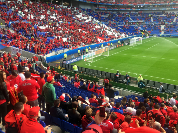 Ein volles Stadion mit Zuschauern, einige sitzen, andere stehen, bei einem Fussballspiel, mit Bannern, einem Torpfosten und Deckenbeleuchtung.