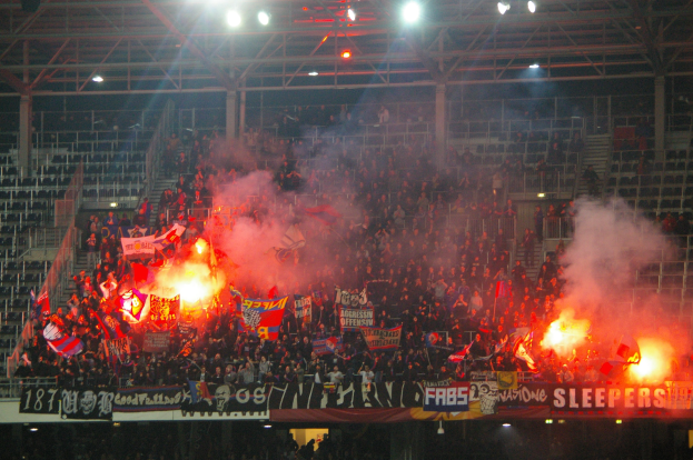 Eine große Menge Menschen in einem Stadion hält Fahnen und Banner, mit Leuchtraketen und Rauch unter einer Decke mit Deckenleuchten und Metallrahmen.