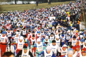 Eine große Gruppe von Menschen, die bei einem Rennen auf einem schneebedeckten Boden laufen, mit Mützen und Skistöcken, umgeben von Bäumen und Text am unteren Bildrand.