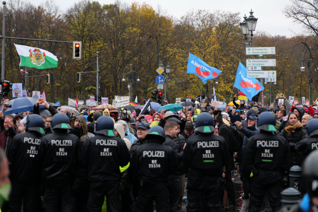 Eine große Gruppe von Polizisten steht vor einer Menge mit Fahnen und Schirmen, mit Laternenmasten, Ampeln, Verkehrszeichen, Bäumen und einem klaren Himmel im Hintergrund.