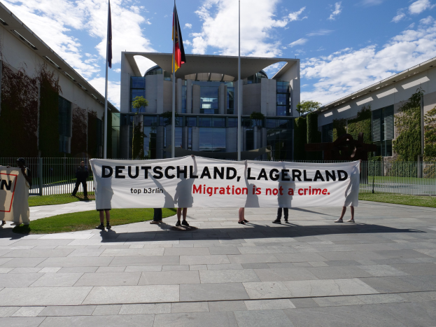 Gruppe von Menschen mit einem Banner "Deutschland, Lagerland Migration ist kein Verbrechen" vor einem umzäunten Gebäude mit Bäumen und Flaggen.