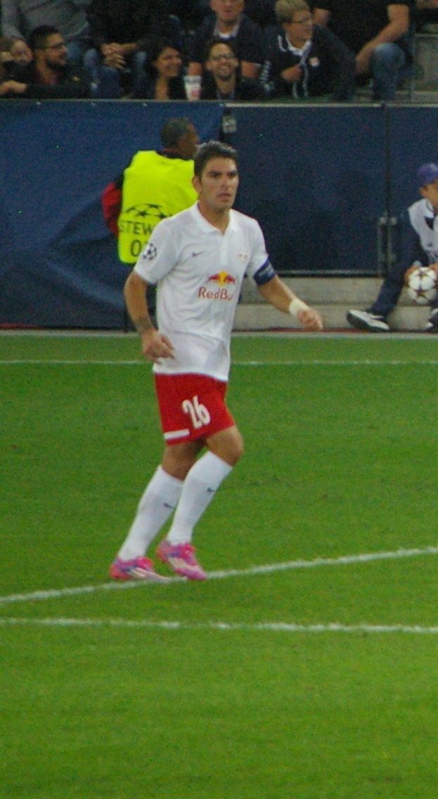 Ein Mann in einem weißen Hemd und roten Shorts, der auf einem Fußballfeld spielt, umgeben von einer Gruppe von Menschen im Hintergrund.