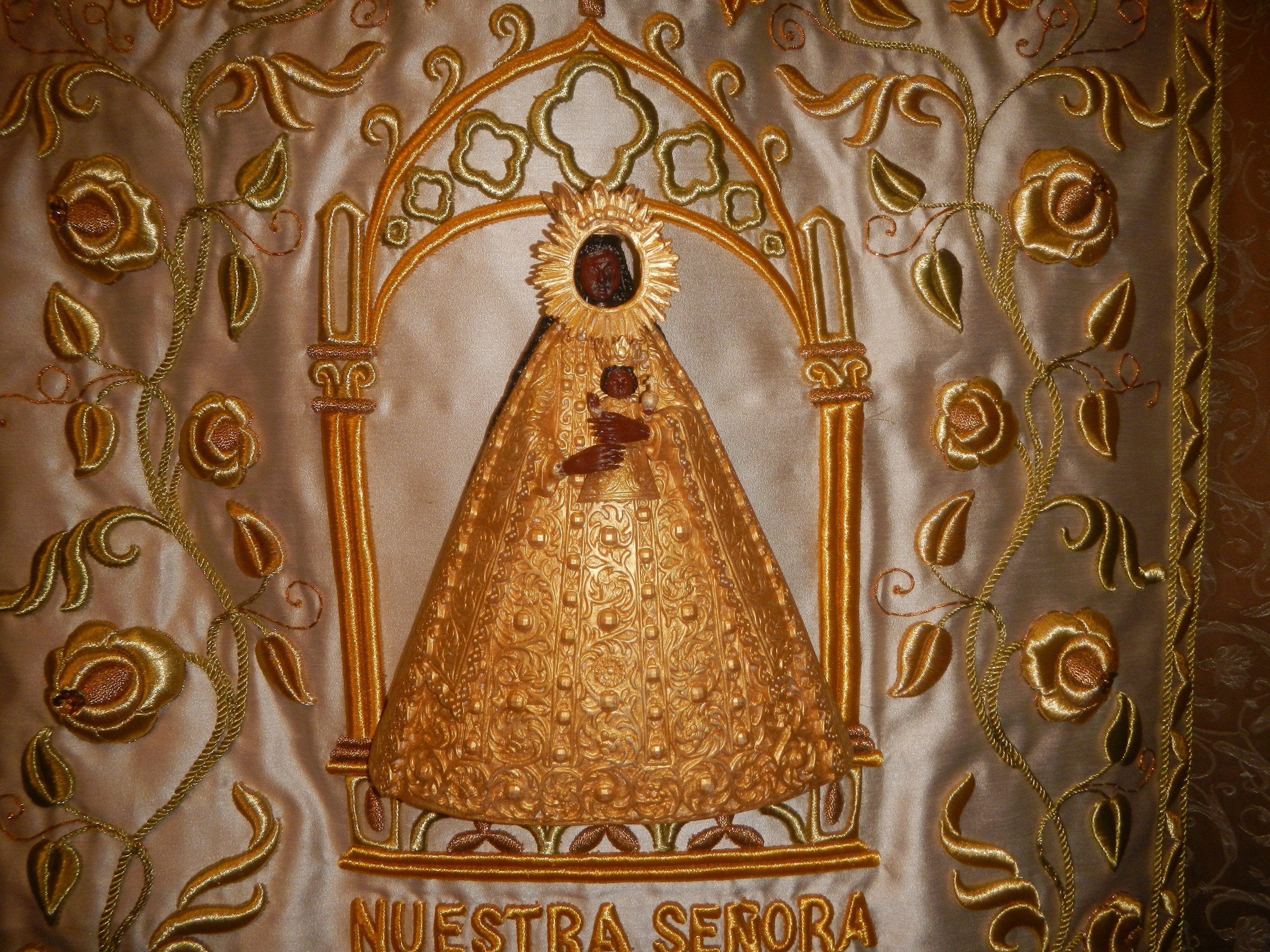 Goldbestickte Chasuble mit einer serenen Frau in einem hochgeschlossenen, langärmeligen Kleid mit einem Dutt, umgeben von kunstvollen Texten und goldenen Verzierungen.