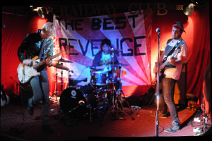 Zwei Personen vor einem Mikrofon stehend und Gitarren spielend, mit einem Mann mit Brille dahinter, der Schlagzeug spielt, vor einem Hintergrund mit hängenden Flexi-Schallplatten und CDs.