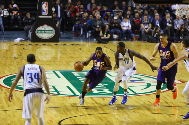 Ein Basketballspiel zwischen den Phoenix Suns und den Dallas Mavericks, mit Spielern auf dem Feld und Zuschauern in den Rängen, mit einem Basketballkorb oben rechts im Bild.