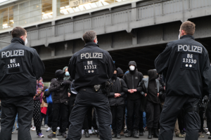 Eine Gruppe von Polizisten in schwarzen Uniformen und Masken steht vor einer Menge, mit einer Brücke und einem Gebäude im Hintergrund, während einer Protest in einer Stadt.