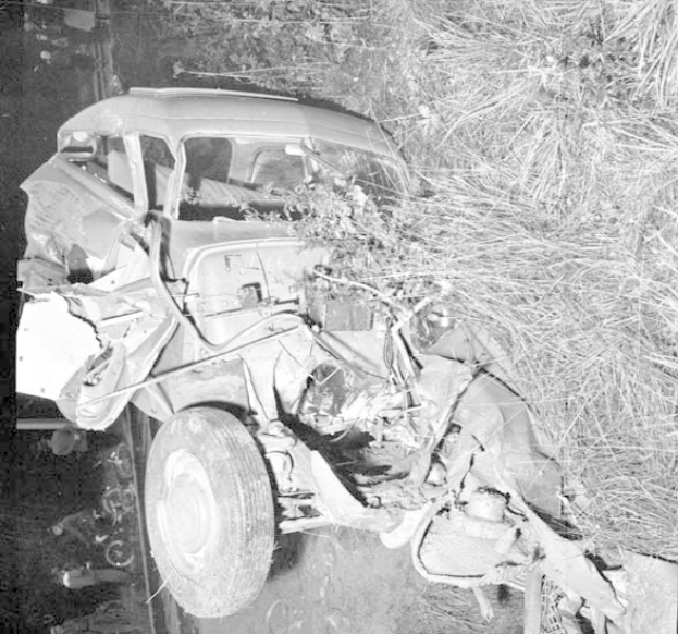 Schwarz-weiß-Foto eines stark beschädigten Autos mit eingedellter Front und zersplitterten Fenstern am Straßenrand, mit einem Baum und verstreuten Gegenständen im Hintergrund.