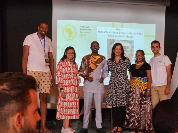 Eine Gruppe von Menschen auf einer Bühne, die für ein Foto posieren und lächeln, mit einem Publikum davor und einem Bildschirm im Hintergrund, auf dem 'Open the Knowledge Journalism Awards 2019' angezeigt wird.