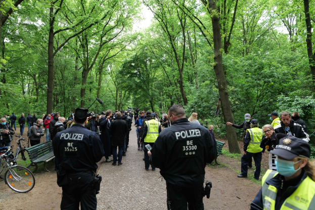 Eine Gruppe von Polizisten steht vor einer Menge von Menschen, einige tragen Mützen und Masken, mit Fahrrädern und einer Bank im Vordergrund und Bäumen und Himmel im Hintergrund.
