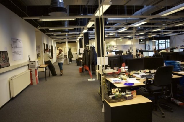 Großes Großraumbüro mit Menschen an Schreibtischen, Computern und Monitoren, umgeben von Papieren, Büchern, Kisten und hängenden Kleidern, mit Text- und Bildtafeln an den Wänden und Deckenleuchten.