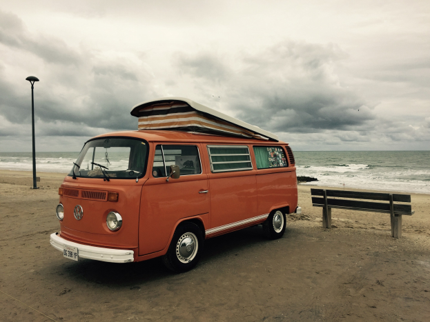 Orange Volkswagen-Bus auf sandigem Strand neben dem Ozean geparkt, mit einer Bank und einem Laternenmast in der N├Ąhe; bew├Âlktes Himmel und sichtbares Wasser im Hintergrund.