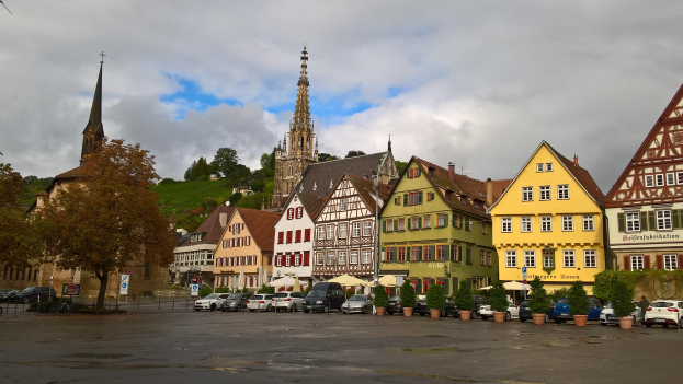 Ein malerischer Marktplatz in Rothenburg ob der Tauber, Deutschland, mit verschiedenen Gebäuden mit Fenstern, Bäumen, Blumentöpfen, Fahrzeugen, Schildern und einer Kirche im Hintergrund bei bewölktem Himmel.