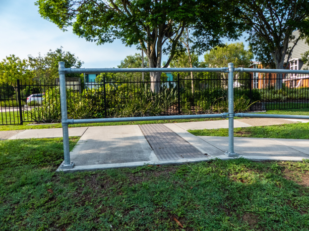 Ein Metallzaun in einer grasbewachsenen Fläche, umgeben von Pflanzen, Bäumen und einem Auto auf der Straße, mit Gebäuden und einem klaren blauen Himmel im Hintergrund.