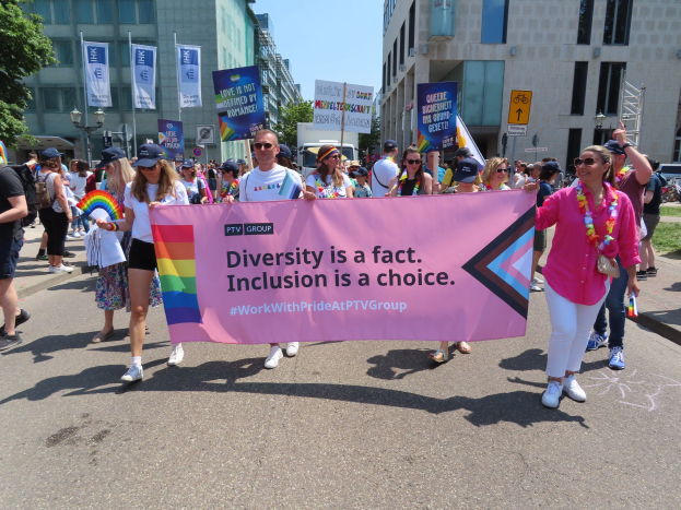 Eine bunte Gruppe von Menschen marschiert auf der Straße, hält ein Banner mit der Aufschrift "Diversität ist eine Tatsache, Inklusion ist eine Wahl", einige tragen Mützen und Schutzbrillen, andere halten zusätzliche Banner, vor einem Hintergrund aus Gebäuden, Bäumen, Laternenpfählen und einem klaren blauen Himmel.