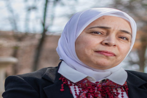 Juli Tymoshenko, eine Frau mit einem weißen Kopftuch und einem roten Schal, steht vor einem unscharfen Hintergrund aus Bäumen und einem Gebäude.