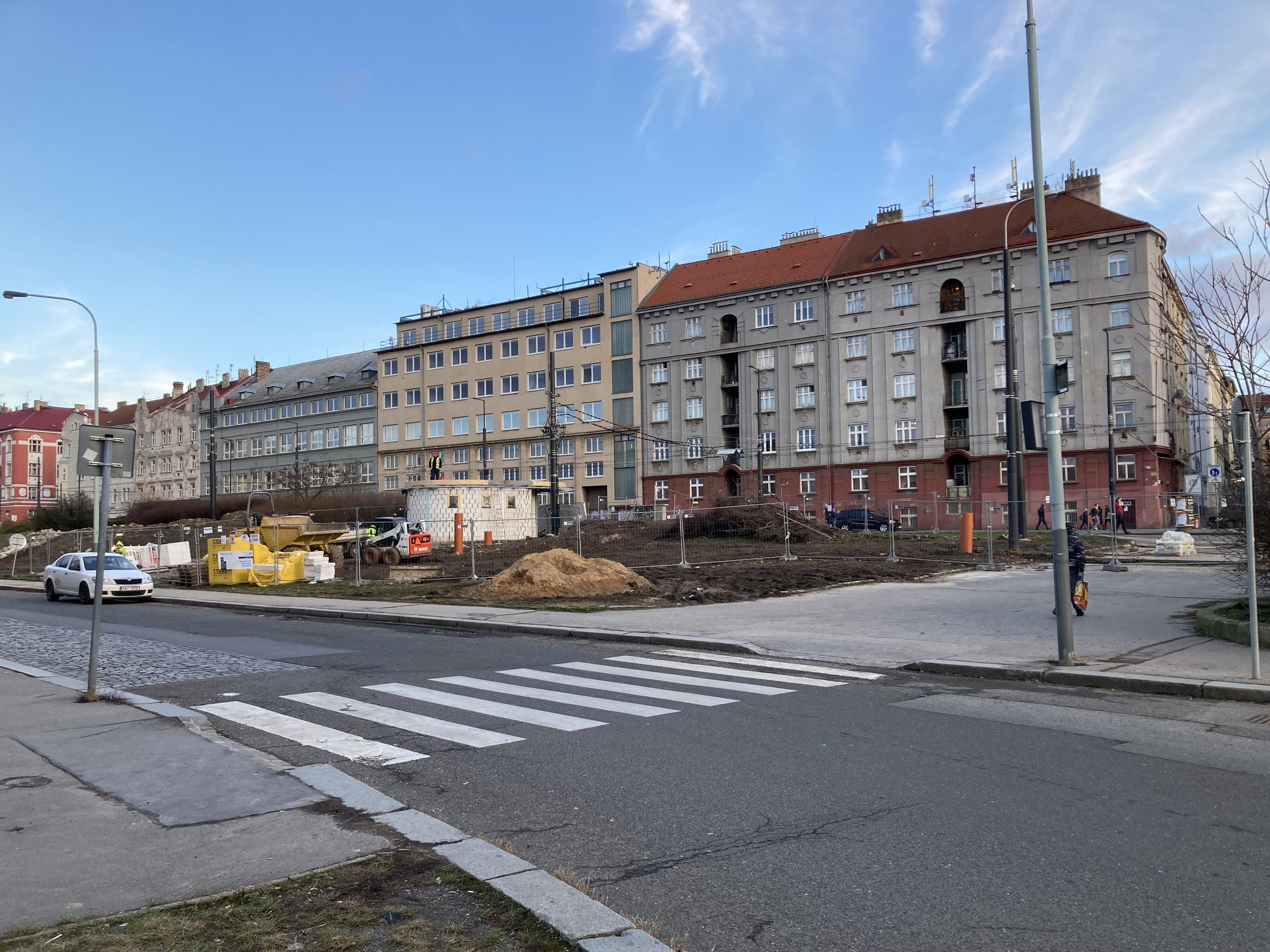 Bauplatz für einen neuen Wohnblock im Vordergrund einer Stadtstraße mit parkenden Autos, Laternen, Bäumen, Gebäuden mit Fenstern und einem bewölkten Himmel im Hintergrund.
