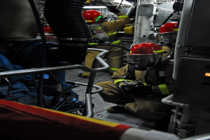 Feuerwehrleute in Helmen und Schutzausrüstung arbeiten an einem Löschfahrzeug mit Rohren und Gegenständen im Hintergrund, unter einer Lampe oben.