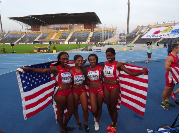 Vier Frauen in Sportbekleidung stehen zusammen auf einer Laufbahn, jede hält eine amerikanische Flagge und lächelt, mit einem Stadion und Zuschauern im Hintergrund.