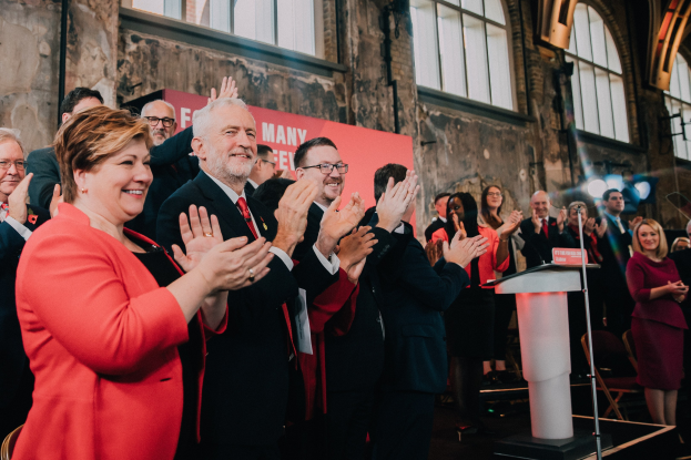 Eine Gruppe von Menschen, die vor einem Publikum applaudieren, mit einem Podium, einem Mikrofon und einer Tafel mit Text auf der rechten Seite und Stühlen, einem Banner, einer Wand, Fenstern und Lichtern im Hintergrund.