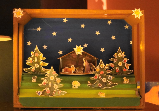 Eine Krippe in einer Holzschachtel auf einem Tisch, dekoriert mit Weihnachtsbäumen, Tieren und einem Haus, mit einer Lampe auf der linken Seite und einer Wand im Hintergrund.