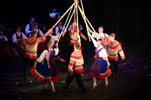 Eine Gruppe von Menschen in farbenfrohen Kostümen tanzt um einen Maibaum, einige halten hölzerne Stäbe, mit Musikern im Hintergrund, die Instrumente spielen.