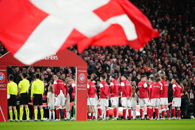 Gruppe von Menschen auf einem Fußballfeld mit einer roten und weißen Flagge im Vordergrund, einem Bogen mit 'Bayern München vs. Bayern München Wetten & Vorschau' im Hintergrund und einer großen Menschenmenge im Stadion.