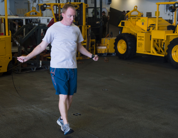 Ein Mann in einem weißen Hemd und blauen Shorts geht durch ein volles Lager, das mit Maschinen, Fahrzeugen, Menschen und diverser Ausrüstung gefüllt ist.
