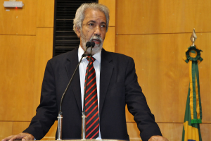 Ein Mann in einem schwarzen Anzug steht an einem Mikrofon und spricht, mit einer braunen Wand und Flaggen im Hintergrund.