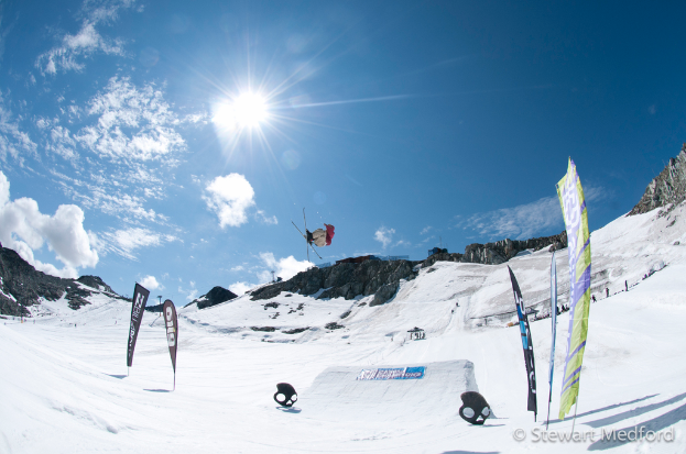 Eine Person in Skiausrüstung ist in der Luft mit schneebedeckten Bergen im Hintergrund.
