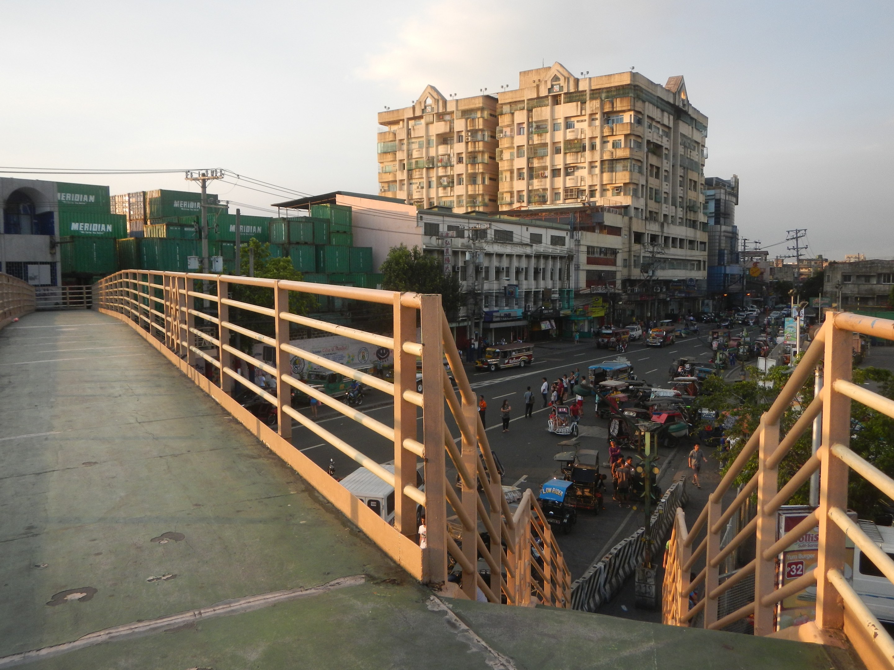Eine Fußgängerbrücke mit Geländern überspannt eine vielbefahrene Straße mit Fahrzeugen und Fußgängern, im Hintergrund sind Gebäude, Bäume und Strommasten zu sehen.