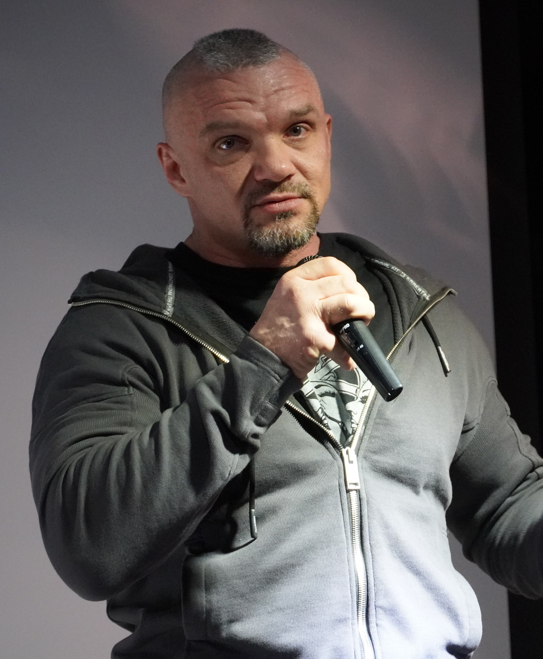 Ein Mann in einer schwarzen Kapuzenjacke und einem schwarzen Anzug mit weißem Hemd hält ein Mikrofon und steht selbstbewusst vor einer Wand, der Mann sieht aus wie ein Komiker mit zurückgekämmtem Haar.
