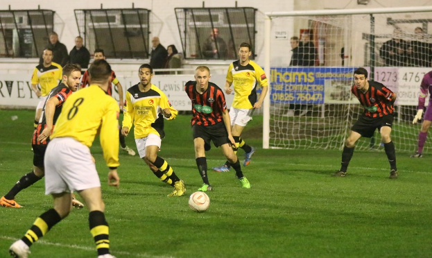 Ein Sportfeld mit mehreren Männern, die Fußball spielen, ein Netz auf der rechten Seite, eine Tafel mit Text im Hintergrund und Zuschauern, die hinter der Tafel stehen, sowie eine Wand und Fenster im Hintergrund.