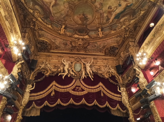 Innensicht eines Theaters mit roten Vorhängen, Kronleuchtern, Skulpturen, Lichtern und einer Deckenmalerei, die eine Bühne mit ein paar Menschen am Boden zeigt.