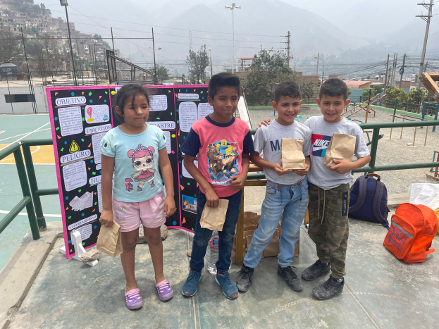 Eine Gruppe von Kindern steht auf einem Basketballfeld, jedes hält einen Papiertüte in der Hand, mit Tüten und Gegenständen auf der rechten Seite, einer Texttafel auf der linken Seite und einer Kulisse aus Zaun, Pfosten, Straßenlaternen, Bäumen, Gebäuden, Hügeln und Himmel.