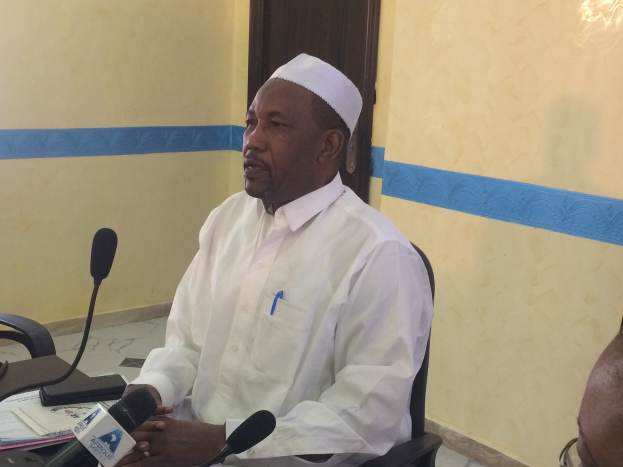 Ein Mann in einem weißen Kleid und Hut sitzt an einem Tisch mit Mikrofonen, spricht, während Papiere und Gegenstände auf dem Tisch sichtbar sind, mit einer anderen Person mit Brille rechts von ihm in einem Raum mit einer Wand und einer Tür.