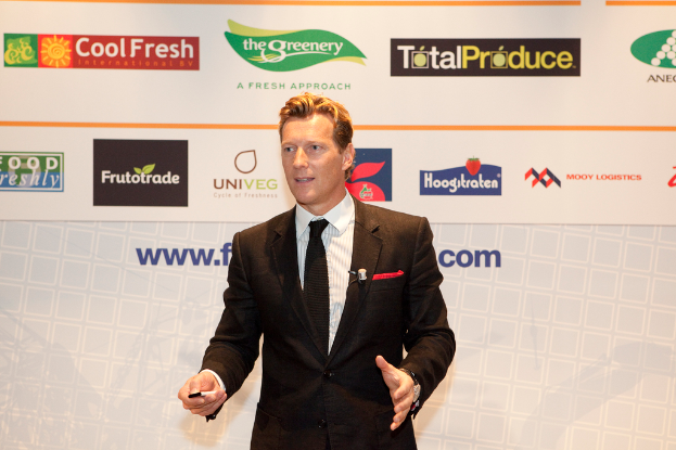 Ein Mann in einem schwarzen Blazer hält ein Objekt und spricht, mit einem weißen Banner und einer weißen Tafel mit Organizational-Logos im Hintergrund.