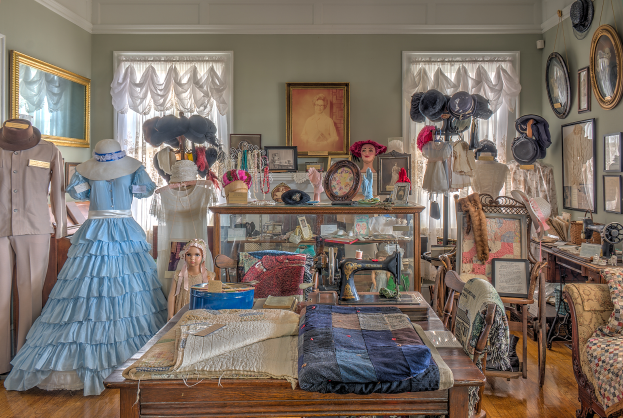 Ein Raum im Museum of Fine Arts in St. Louis, Missouri, gefüllt mit Puppen in Kleidern und Hüten, einem Tisch mit Kleidung und einer Nähmaschine, Stühlen mit Kissen, einem Schrank mit verschiedenen Gegenständen, Fotorahmen an der Wand, Fenstern mit Vorhängen und einer Decke.