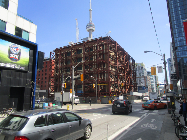 Eine belebte Stadtstraße mit fahrenden Autos und Fahrrädern, Fußgängern auf dem Gehweg, einem großen Gebäude im Hintergrund, das gebaut wird, städtische Infrastruktur einschließlich Masten, Lampen, Verkehrszeichen und Schilder, mit dem CN Tower in der Ferne unter einem klaren blauen Himmel.