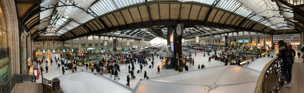 Eine große Gruppe von Menschen mit Gepäck geht durch einen hell erleuchteten Bahnhof, mit einer Uhr und Textanzeigen im Hintergrund.