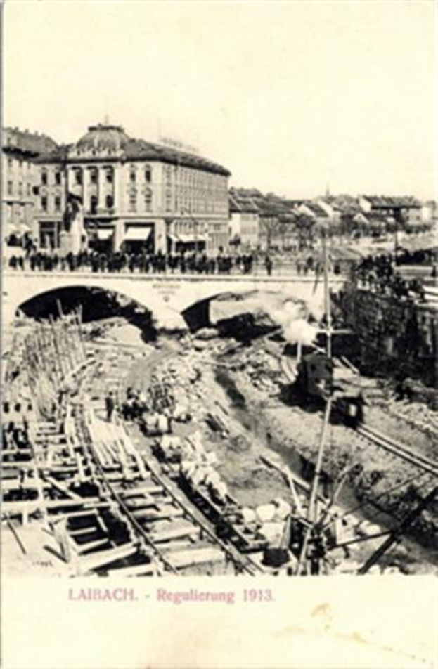 Eine Schwarz-Weiß-Postkarte aus dem Jahr 1913, die eine Bahntrasse mit einer Brücke im Hintergrund zeigt, umgeben von Gebäuden, Menschen, Pfählen, Bäumen und anderen Gegenständen, mit Text am unteren Rand, der den Bau der Bahntrasse in Laibach anzeigt.