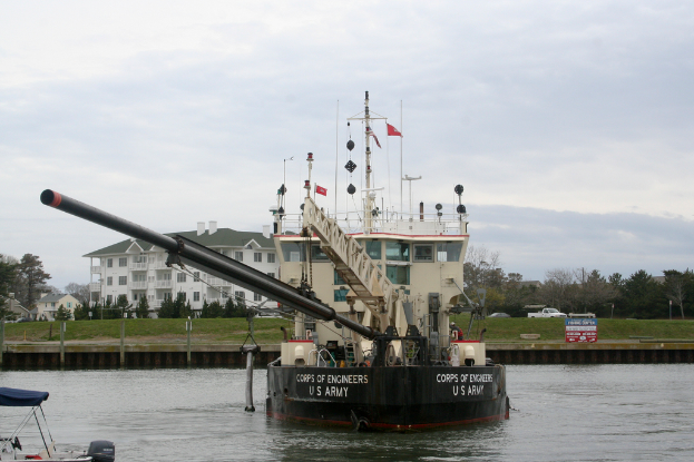 Ein Schiff auf dem Wasser mit Bäumen, Fahrzeugen, Gebäuden und Polen im Vordergrund und einem Himmel im Hintergrund, mit einem Boot in der linken unteren Ecke.
