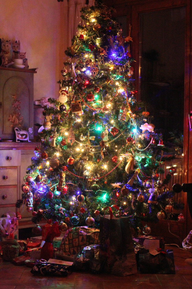 Ein Weihnachtsbaum mit Lichtern und Dekorationen, Geschenke auf dem Boden, ein Schrank mit Gegenständen links und ein Fenster an der rechten Wand.