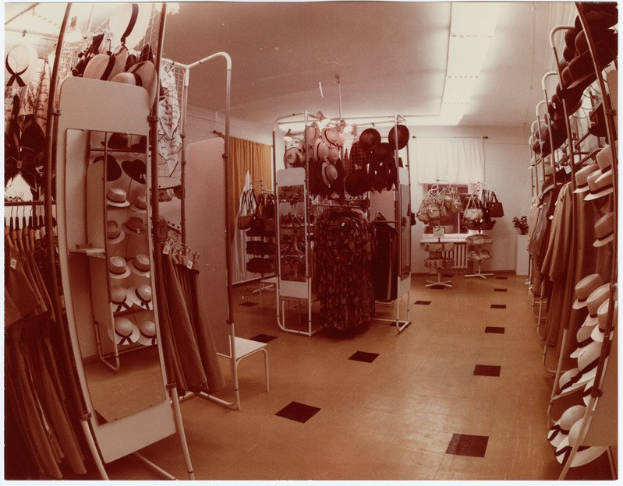 Altes Schwarz-Weiß-Foto eines kleinen Kleiderladens mit Kleiderstangen, Hutständern, einem Spiegel, einem Stuhl, einem Tisch mit diversen Gegenständen, gemusterten Vorhängen und Deckenbeleuchtung.