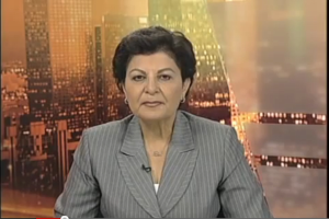 Eine Frau in einem Business-Outfit sitzt vor einem Fernseher mit Text am unteren Bildschirmrand, mit ein paar Gebäuden im Hintergrund.