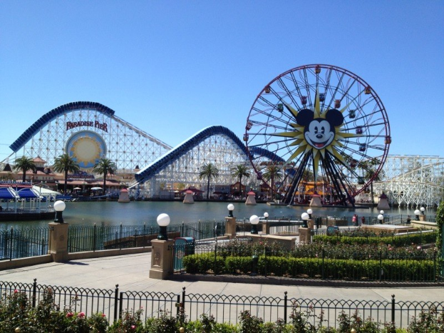 Luftaufnahme von Disney California Adventure mit seinem Riesenrad und Achterbahn, umgeben von Grünflächen und Laternen, mit Wasser im Vordergrund und einem klaren blauen Himmel.