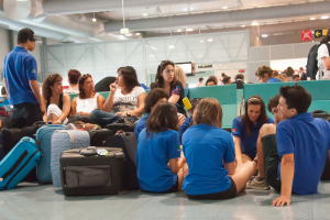 Ein belebtes Flughafeninnere mit vielen Menschen, die auf Flüge warten, Gepäck auf der linken Seite und Glastüren mit Richtungsschildern im Hintergrund.