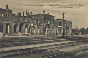 Schwarz-weiß-Fotografie des Bahnhofs Korschen in Ostern, Deutschland, mit Bahnschienen, Bahnhofsgebäuden, Menschen, Bäumen und Himmel.